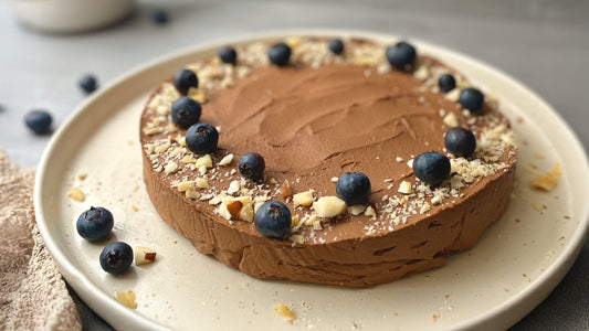 Ein runder, cremiger Schokokuchen auf einem hellen Teller. Der Kuchen ist mit frischen Heidelbeeren und gehackten Paranüssen am Rand dekoriert. Der Hintergrund ist weich fokussiert, mit vereinzelten Beeren und einem beigefarbenen Tuch am Bildrand.