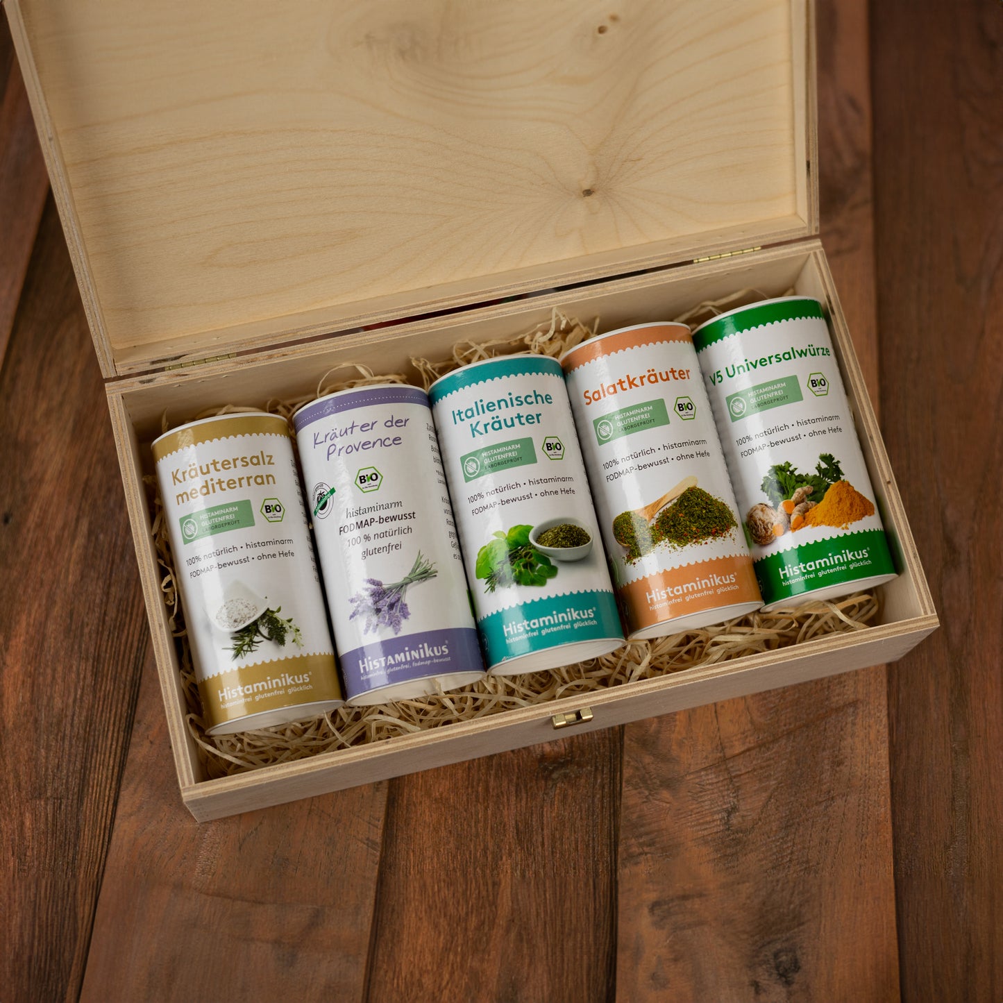 Offene Holzbox auf Holztisch, gefüllt mit fünf Gewürzdosen auf Holzwolle. Sichtbare Sorten: „Kräutersalz mediterran“, „Kräuter der Provence“, „Italienische Kräuter“, „Salatkräuter“ und „V5 Universalwürze“. Jede Dose mit Bio-Siegel und farbigem Etikett.