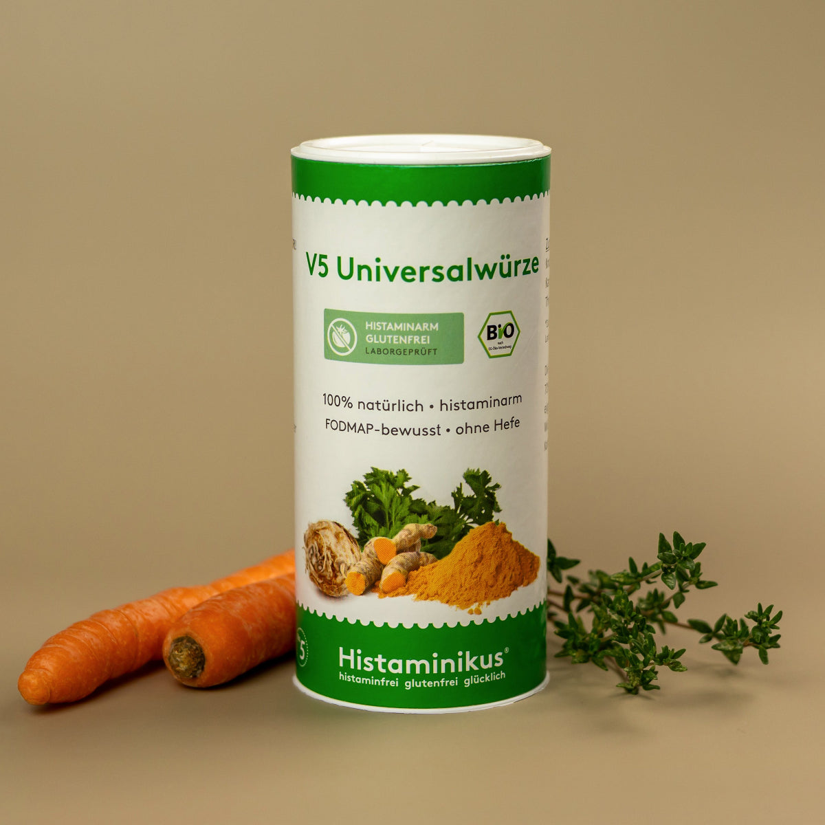 Dose der V5 Universalwürze von Histaminikus auf beigem Untergrund, daneben zwei Karotten und ein Zweig Thymian. Auf der Dose abgebildet sind Petersilie, Knollensellerie, Kurkuma und weitere Gewürze. Aufdrucke: „histaminarm“, „glutenfrei“, „FODMAP-bewusst“, „ohne Hefe“ und „Bio“.