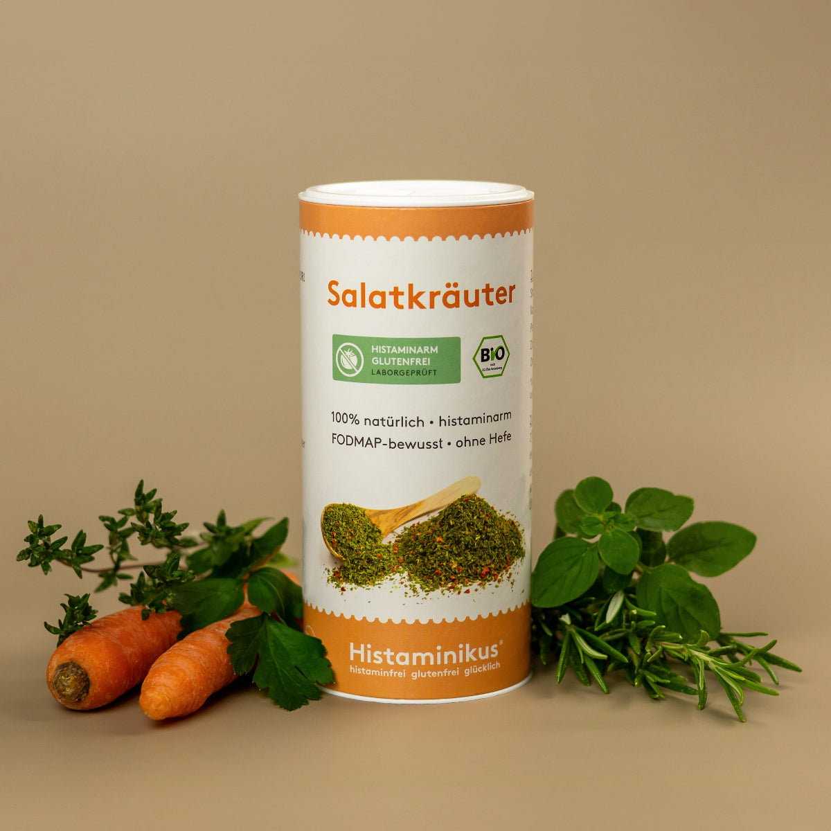 Foto der Gewürzdose „Salatkräuter“ von Histaminikus mit natürlicher Dekoration. Auf dem Etikett stehen Hinweise wie „100 % natürlich“, „histaminarm“, „FODMAP-bewusst“ und „ohne Hefe“. Unten Markenlogo mit Claim. Im Vordergrund frische Karotten, Kräuter und grüne Blätter.