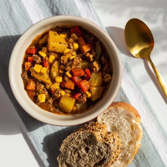 Schale mit herzhaftem No!Chili con Carne, gefüllt mit Paprika, Kartoffeln, Mais, Karotten und veganem Hack, serviert auf einem hellen Tuch mit goldfarbener Löffelgarnitur und zwei Brotscheiben im Vordergrund.