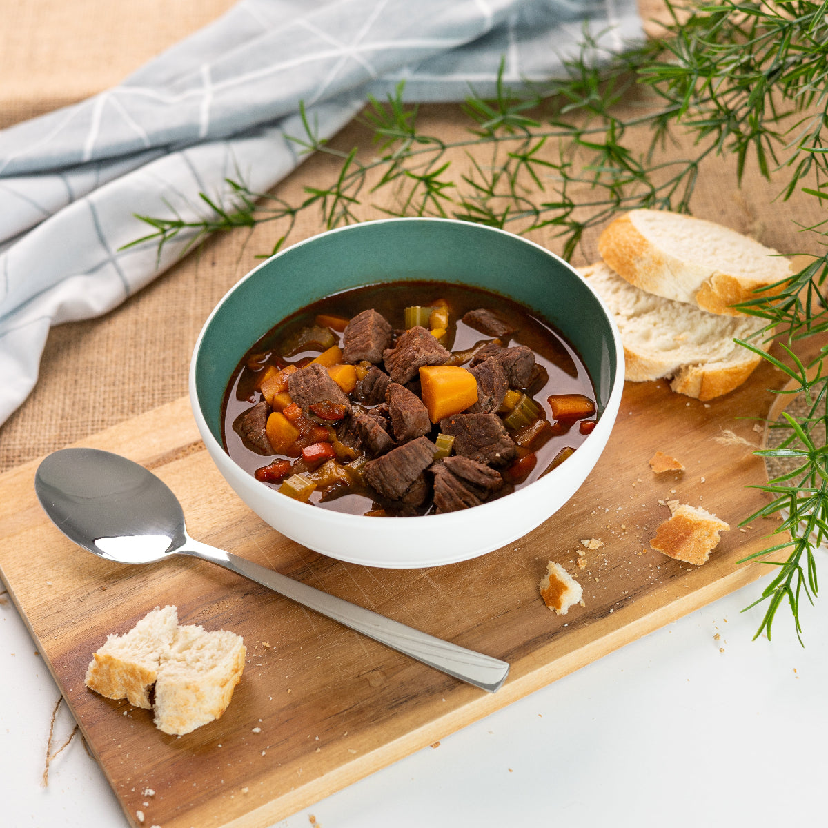 Eine Schale mit saftigem Gulasch, angerichtet mit würfeligem Rindfleisch, buntem Gemüse und kräftiger Soße. Daneben frisches Baguette, ein Löffel und ein paar Brotkrümel auf einem Holzbrett – gemütlich angerichtet mit Leinentuch und Rosmarinzweig im Hintergrund.