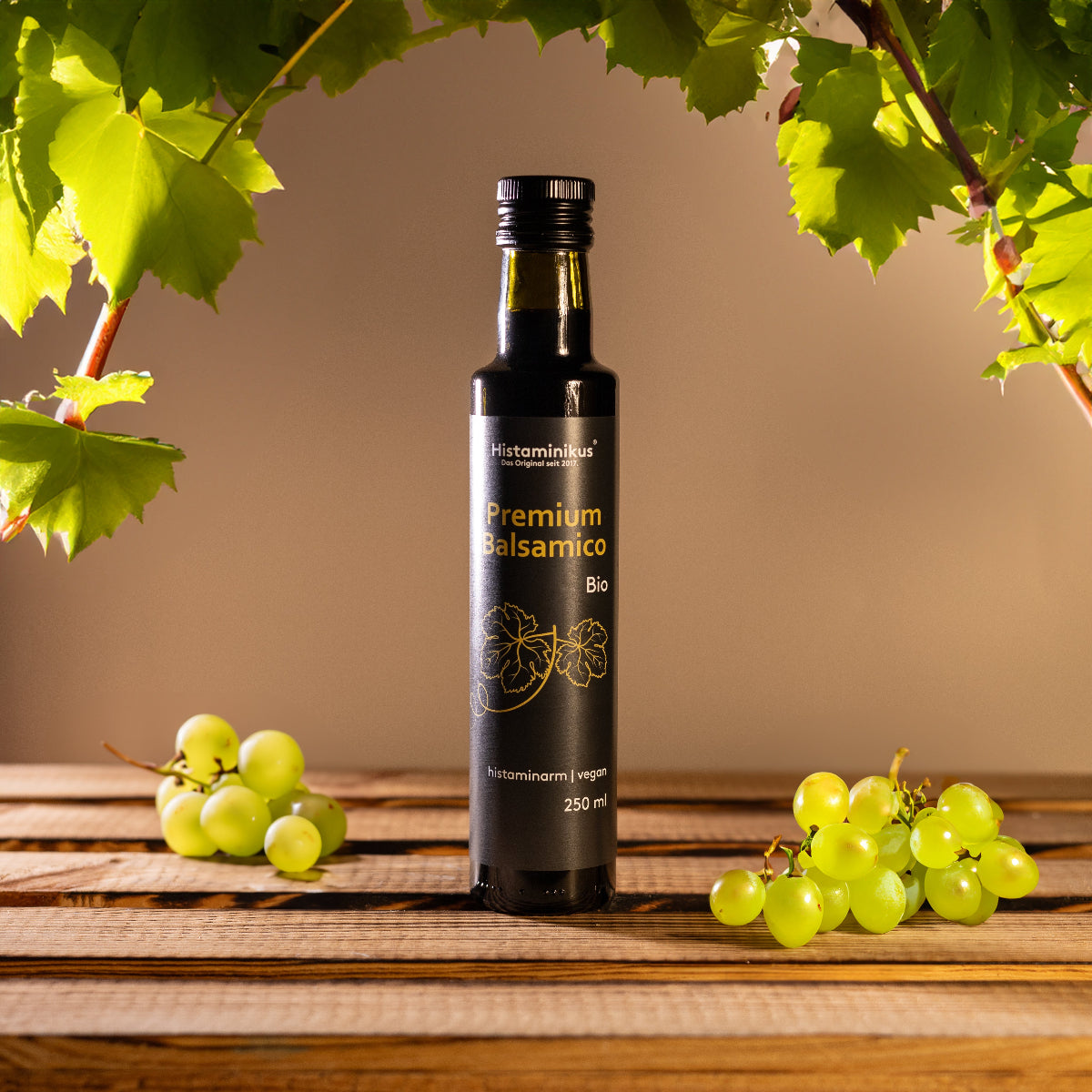 Eine schwarze Flasche „Premium Balsamico Bio“ von Histaminikus steht auf einem Holztisch, umgeben von grünen Weintrauben. Im Hintergrund hängen grüne Weinblätter herab. Die Flasche ist als histaminarm und vegan gekennzeichnet.