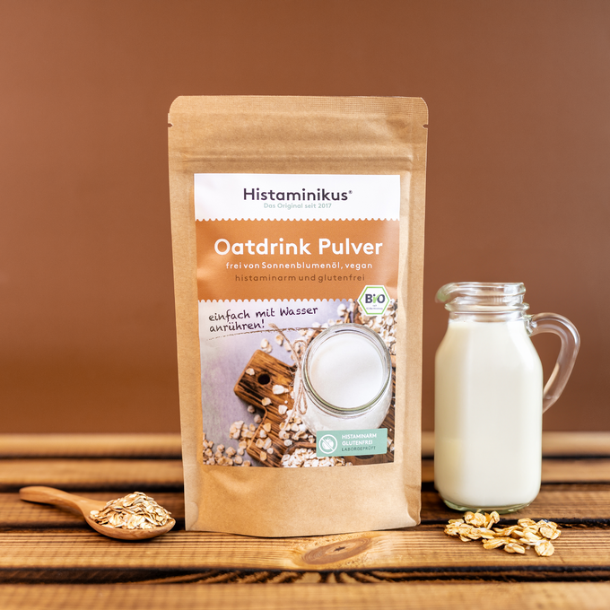 Oatdrink Pulver von Histaminikus in braunem Beutel, daneben eine Glaskaraffe mit angerührtem Haferdrink und ein Holzlöffel mit Haferflocken auf Holztisch vor braunem Hintergrund.