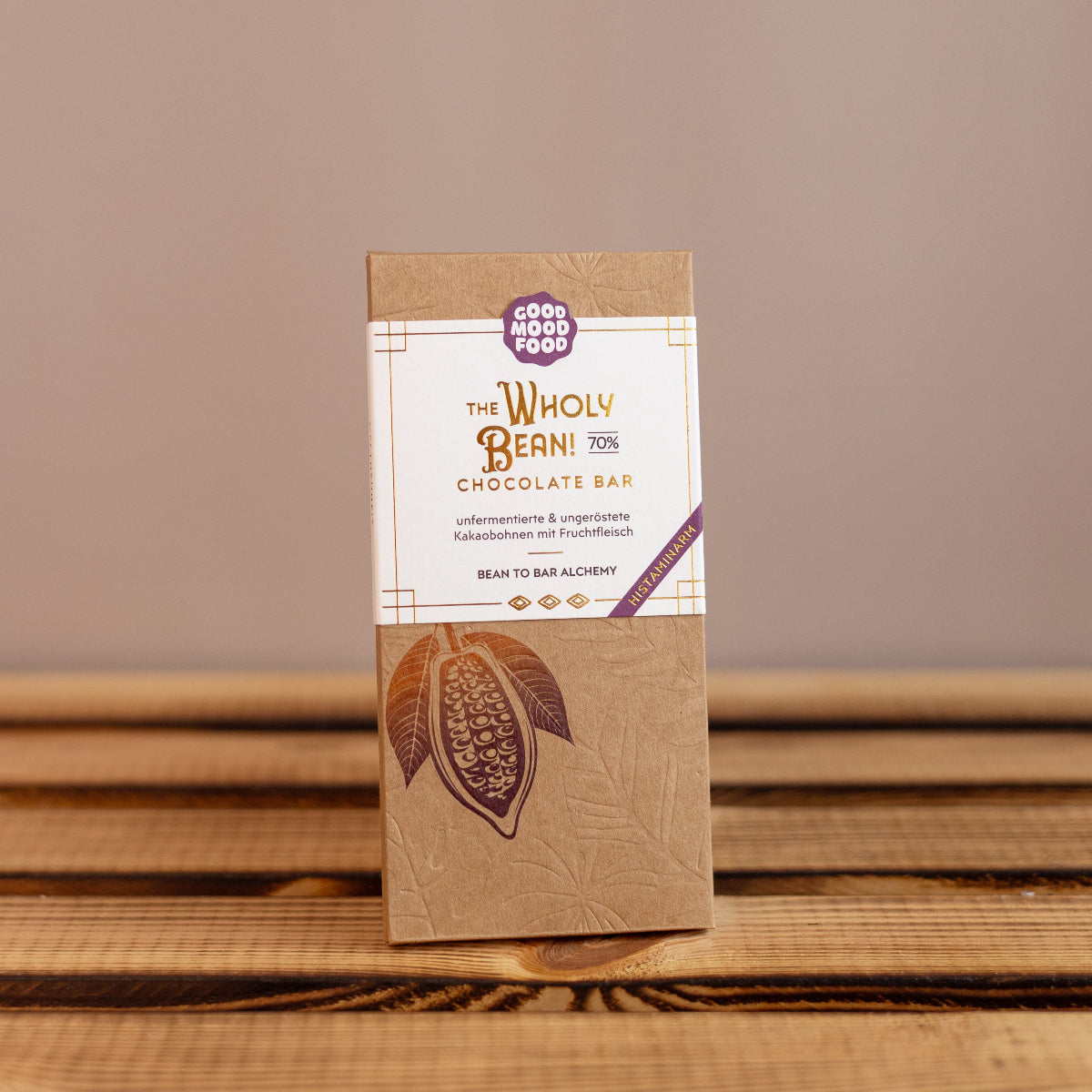 Verpackte Schokolade von Good Mood Food mit der Aufschrift ‚The Wholy Bean! 70 % Chocolate Bar‘ auf einem Holztisch. Die Verpackung ist braun mit weißem Etikett und zeigt eine stilisierte Kakaofrucht.