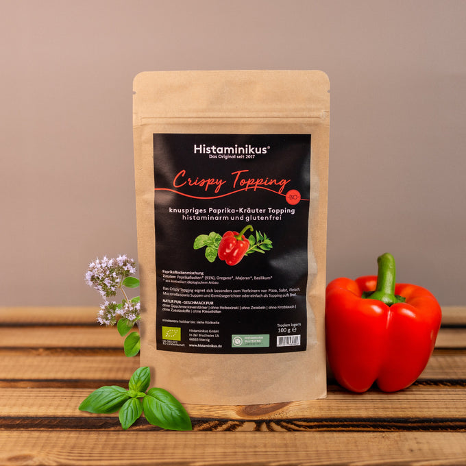 Produktfoto: Braune Standbodenverpackung „Crispy Topping“ mit schwarzem Etikett auf Holzuntergrund. Daneben frische Kräuter (Basilikum, Majoran) und eine rote Paprika. Hintergrund neutral beige.