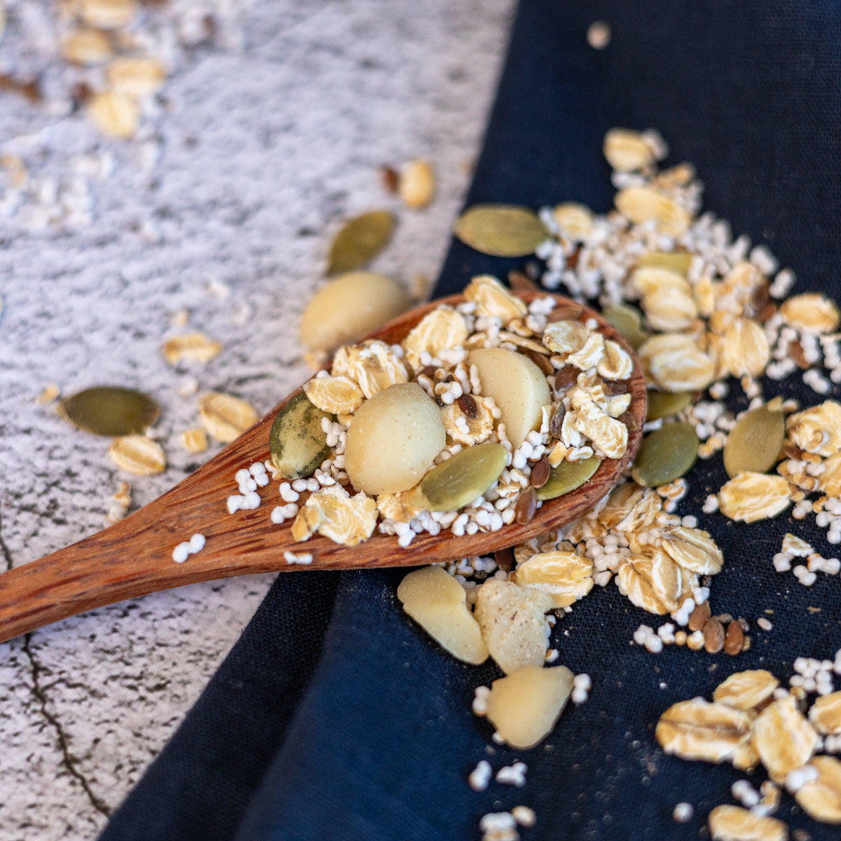 Nahaufnahme eines Holzlöffels, der mit Knuspermüsli gefüllt ist. Zu erkennen sind glutenfreie Haferflocken, Macadamianüsse, Kürbiskerne, gepuffter Amaranth, Leinsamen und Chiasamen. Die Müslimischung ist auf einem hellen Untergrund sowie einem dunklen Tuch verstreut.