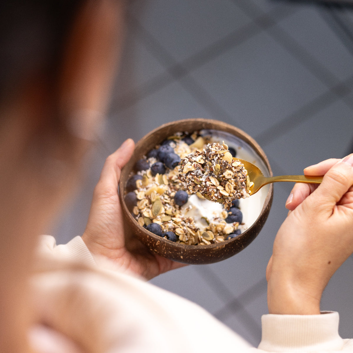 Eine Person aus der Perspektive von hinten beim Frühstücken. Sie hält eine Kokosschale mit Joghurt, Müsli und Heidelbeeren in der linken Hand, während sie mit der rechten einen goldfarbenen Löffel zum Mund führt. Auf dem Löffel liegt eine Portion Knuspermüsli, bei der die Körner und Nüsse gut zu erkennen sind.