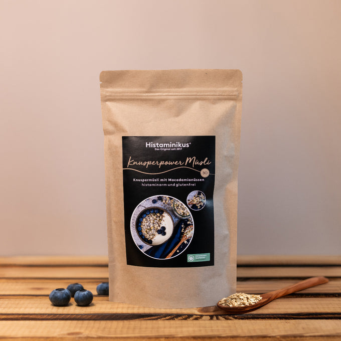Braune Papierverpackung mit schwarzem Etikett. Aufschrift: „Histaminikus – Knusperpower Müsli“. Darunter: „Knuspermüsli mit Macadamianüssen, histaminarm und glutenfrei“. Mittig ein rundes Foto mit Müsli, Joghurt und Blaubeeren in einer Schale sowie eine kleinere Nahaufnahme des Müslis. Im Vordergrund liegen einige Blaubeeren und ein Holzlöffel mit Haferflocken auf einem Holztisch.