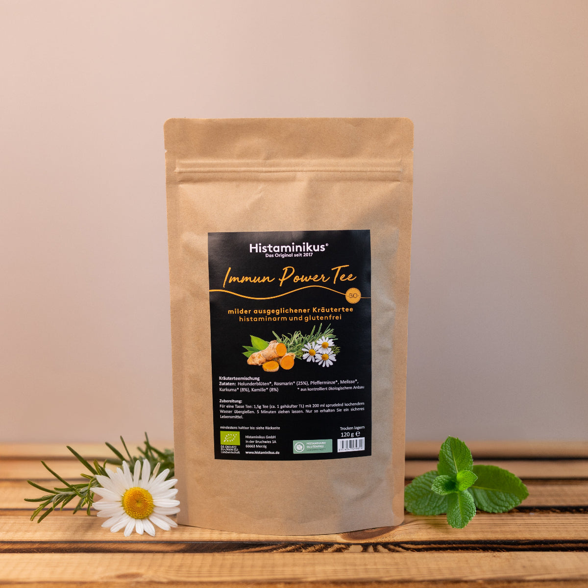 Standbeutel „Immun Power Tee“ von Histaminikus auf einem Holztisch, daneben frische Kamillenblüte, Rosmarin und Minze. Schwarzes Etikett mit orangener Schrift, Aufschrift: „milder ausgeglichener Kräutertee – histaminarm und glutenfrei“. Bio-Siegel und Zutatenillustration.