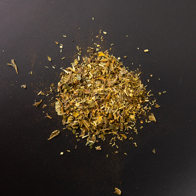 Loser Kräutertee in goldgrüner Farbgebung auf schwarzem Untergrund, Mischung mit erkennbaren Blatt-, Blüten- und Stängelanteilen, darunter feinere gelbliche und bräunliche Bestandteile.