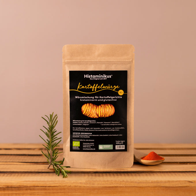Braune Packung „Kartoffelwürze“ von Histaminikus steht auf einem Holzuntergrund vor hellbraunem Hintergrund. Daneben ein Rosmarinzweig und ein Holzlöffel mit rotem Gewürzpulver. Die Würzmischung ist histaminarm, glutenfrei und bio-zertifiziert.