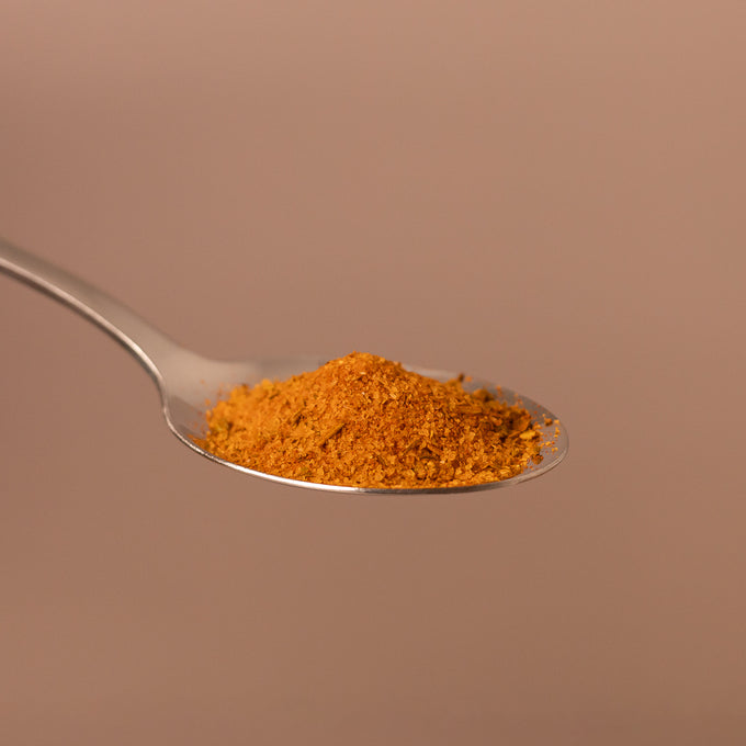 Silberner Löffel mit einem gehäuften Haufen fein gemahlener, orange-brauner Gewürzmischung vor einfarbigem Hintergrund.