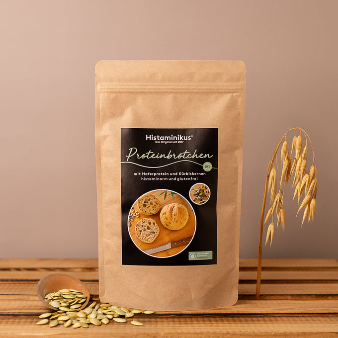 Braune Papiertüte mit schwarzem Produktetikett „Histaminikus Proteinbrötchen“ mit Foto von den Brötchen, neben der Tüte ein Holzlöffel mit Kürbiskernen und Haferhalm-Deko auf Holzuntergrund.