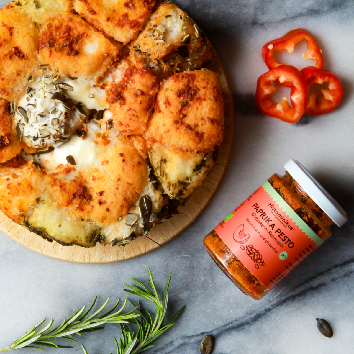 Glas des Paprika Pestos auf eine Mamorplatte. Daneben ein rustikales, rundes Brot mit Körnern und Kräutern. Als Deko liegen noch Rosmarinzweige und drei rote Paprikaringe, sowie ein paar Kürbiskerne dabei.