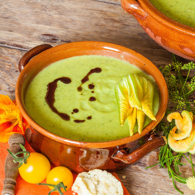 Eine braune Keramikschale, gefüllt mit grüner Zucchinisuppe, garniert mit Zucchiniblüten und einem Muster aus dunklem Öl. Die Schale steht auf einem rustikalen Holztisch, umgeben von einem Stück Brot, einem Holzlöffel und orangefarbenen Kapuzinerkresseblüten.