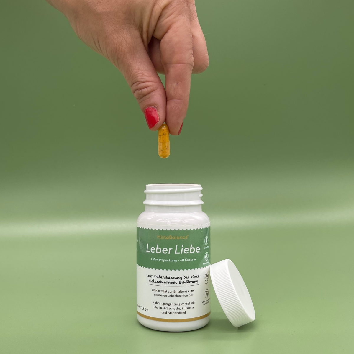 Geöffnete Dose „Leber Liebe“ mit abgenommenem Deckel auf grünem Hintergrund. Eine Hand mit rotem Nagellack hält eine gelbe Kapsel über die Dose. Auf dem Etikett: „zur Unterstützung bei einer histaminarmen Ernährung“ mit Cholin, Artischocke, Kurkuma und Mariendistel.