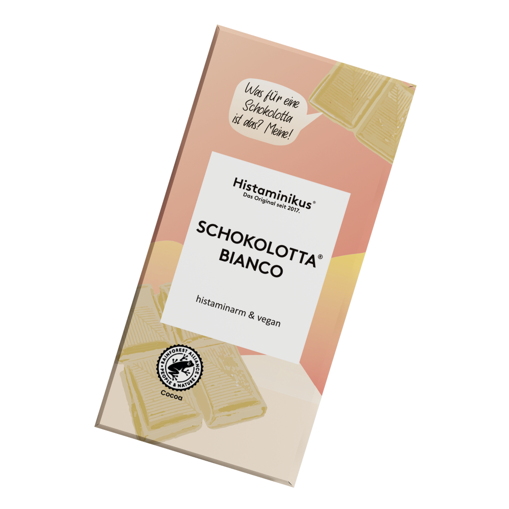 Verpackung der weißen Schokolade „Schokolotta® Bianco“ von Histaminikus in schräger Ansicht. Das Design kombiniert zartes Rosa, Gelb und Beige mit weißen Akzenten. Aufdrucke betonen die Eigenschaften „histaminarm & vegan“ sowie „aus kontrolliertem Anbau“. Im oberen Bereich eine Sprechblase mit dem Text: „Was für eine Schokolotta ist das? Meine!“. Am unteren Rand eine stilisierte Kakaobohne mit dem Hinweis auf nachhaltigen Anbau.