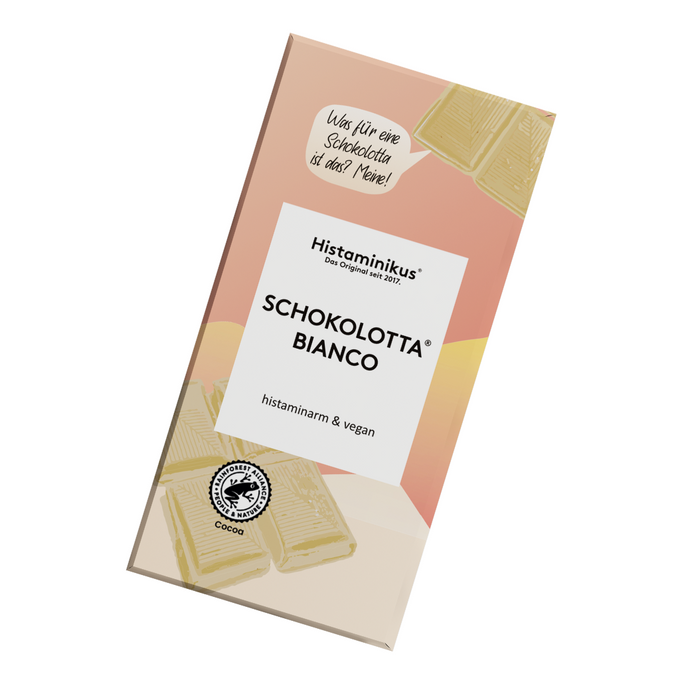 Verpackung der weißen Schokolade „Schokolotta® Bianco“ von Histaminikus in schräger Ansicht. Das Design kombiniert zartes Rosa, Gelb und Beige mit weißen Akzenten. Aufdrucke betonen die Eigenschaften „histaminarm & vegan“ sowie „aus kontrolliertem Anbau“. Im oberen Bereich eine Sprechblase mit dem Text: „Was für eine Schokolotta ist das? Meine!“. Am unteren Rand eine stilisierte Kakaobohne mit dem Hinweis auf nachhaltigen Anbau.