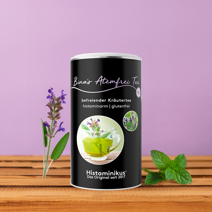 Produktdose „Bina’s Atemfrei Tee“ auf Holzoberfläche vor fliederfarbenem Hintergrund. Links eine frische, blühende Kräuterstiel mit Blättern, rechts ein kleiner Zweig frischer Minze. Auf dem schwarzen Etikett steht „befreiender Kräutertee – histaminarm | glutenfrei“ mit Foto einer Glas-Tasse mit grünem Kräutertee und Kräutern.