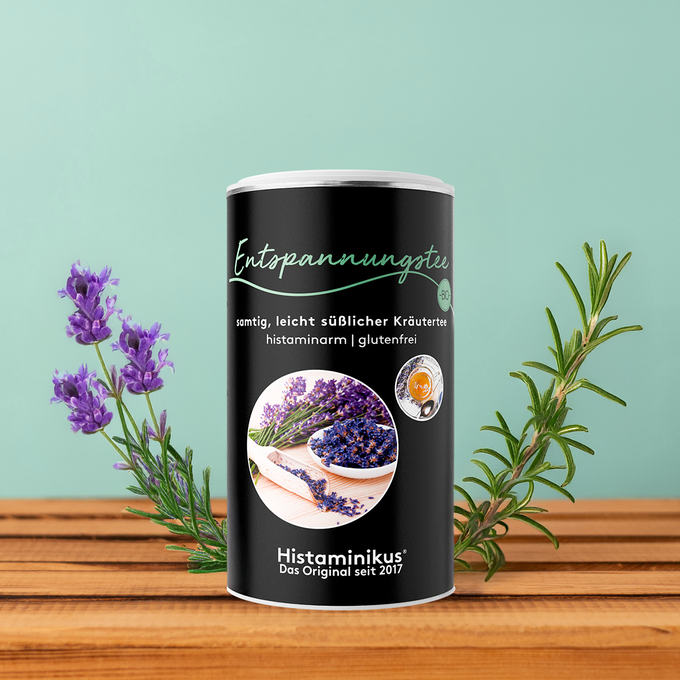 Schwarze Teedose „Entspannungstee“ von Histaminikus auf Holzoberfläche vor mintgrünem Hintergrund, dekoriert mit Lavendel- und Rosmarinzweigen.