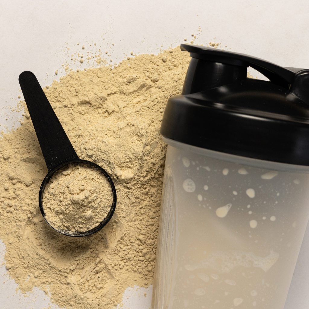 Nahaufnahme der Histaminikus „Proteinquelle“-Mischung: Ein schwarzer Messlöffel liegt auf einer Portion Proteinpulver, daneben ein transparenter Shaker mit schwarzem Deckel.