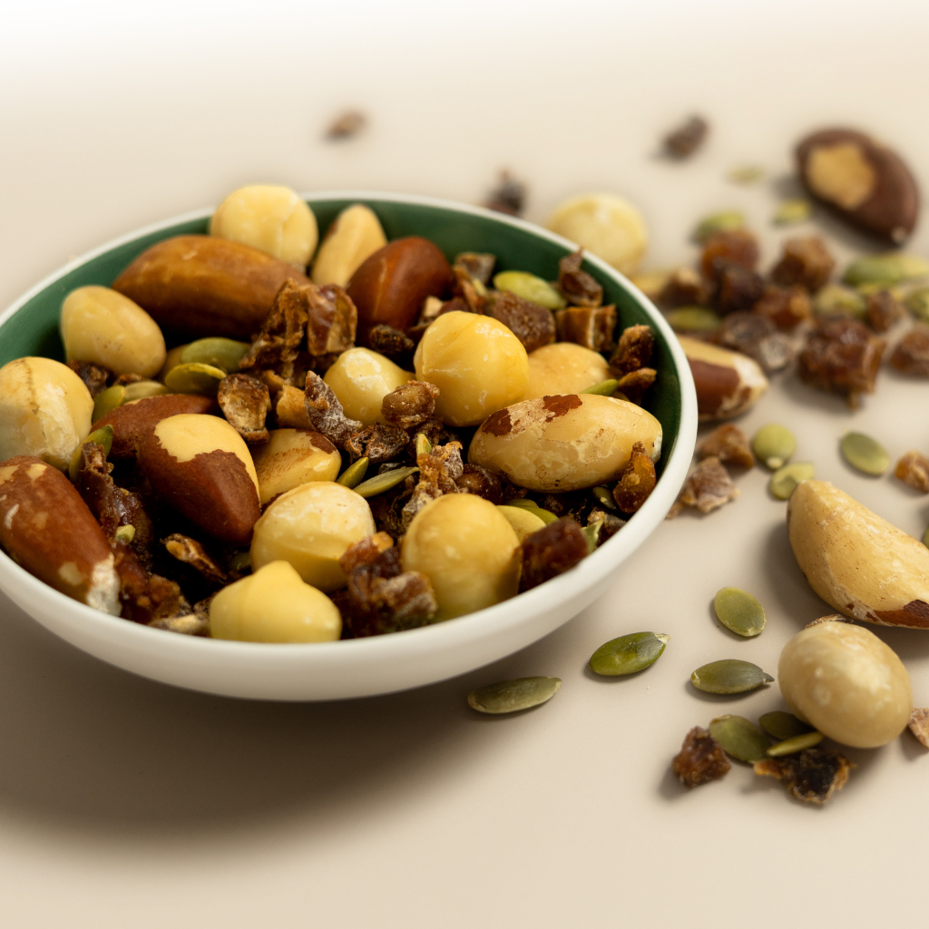 Schale mit Nuss-Frucht-Mischung auf hellem Untergrund.
Enthalten sind Paranüsse, Macadamias, Cashews, Kürbiskerne und kleine Fruchtstücke.
Einige Zutaten liegen auch dekorativ neben der Schale.
Fokus auf Textur und Frische der einzelnen Komponenten.
