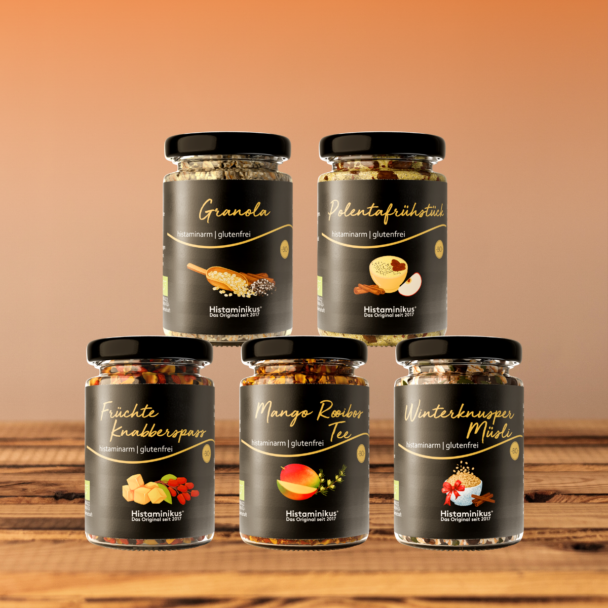 Fünf histaminarme, glutenfreie Frühstücksprodukte von Histaminikus in Gläsern: Granola, Polentafrühstück, Früchte Knabberspaß, Mango Rooibos Tee und Winterknusper Müsli, arrangiert auf einem Holztisch vor warmem Farbverlauf-Hintergrund.