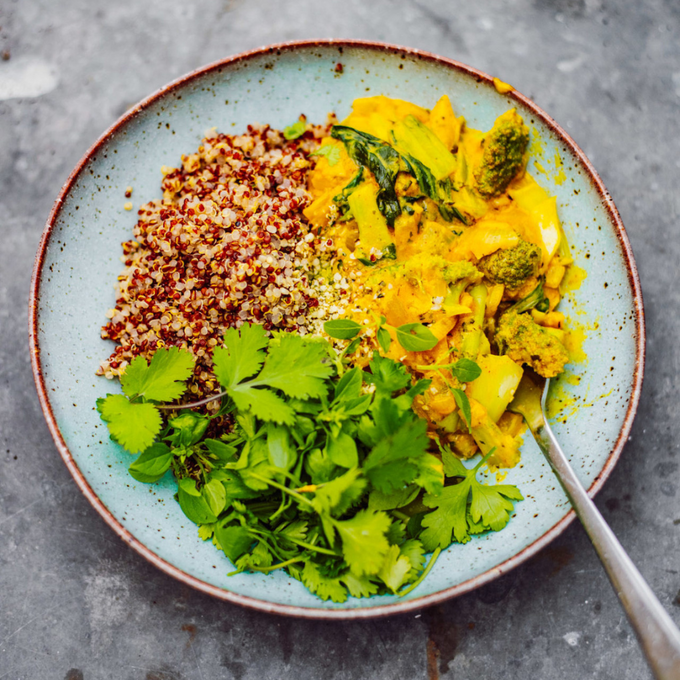 Bunter Teller mit Quinoa-Mix, gelbem Curry mit Gemüse und frischem Koriander. Serviert in einer hellblauen Schale mit braunem Rand, darunter grauer Steinuntergrund. Ein Löffel liegt am rechten Schüsselrand.
