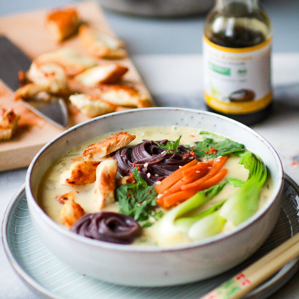 Bunte Bowl mit heller Suppe, violetten Nudeln, Hähnchenstücken, Karotten, Pak Choi und Kräutern. Im Hintergrund Brett mit geschnittenem Hähnchen und Flasche Coco Aminos.