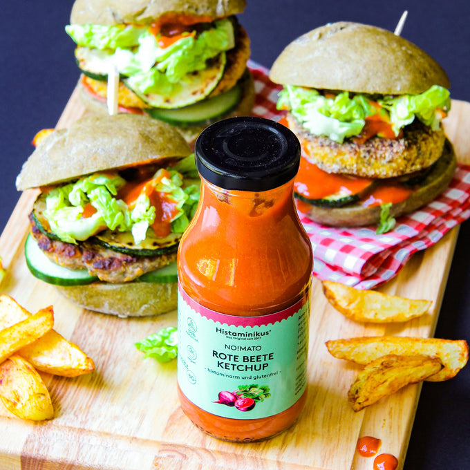 Foto mit der Flasche des Rote Beete Ketchup im Vordergrund. Hinter der Flasche liegen 3 Burger, die mit dem Ketchup zubereitet sind. Neben der Flasche und den Burgern liegen Kartoffelspalten. Alles zusammen ist auf einem Holztablett angerichtet.