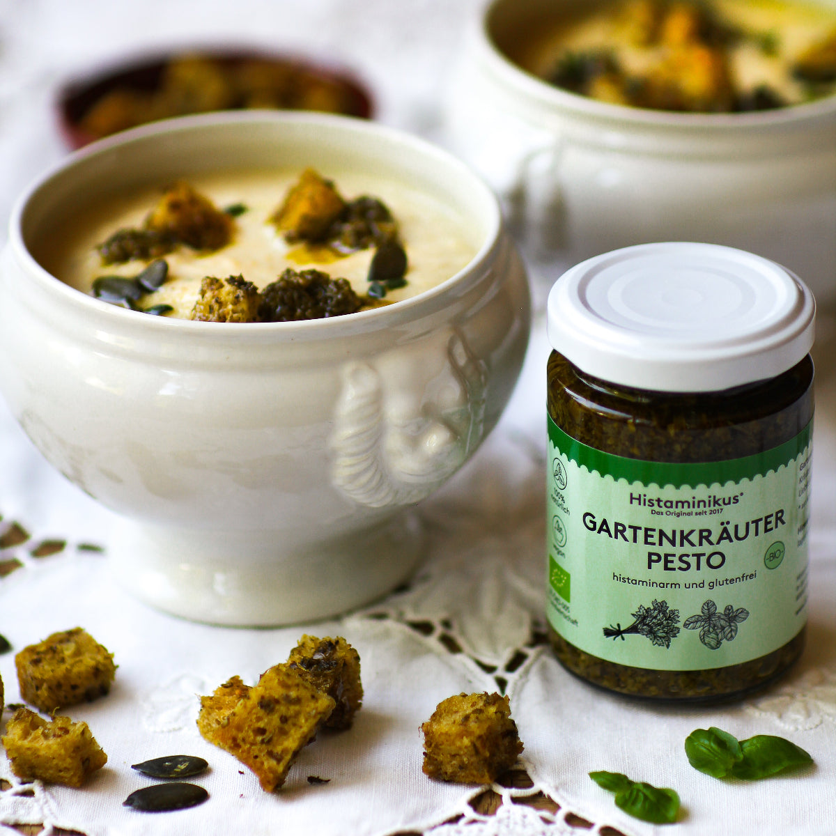 Cremesuppe in einer weißen Schale, garniert mit Croutons, Kürbiskernen und grünem Pesto. Daneben ein Glas Histaminikus Gartenkräuter Pesto mit Bio-Siegel, beschriftet als histaminarm und glutenfrei. Umgeben von weiteren Croutons und frischen Basilikumblättern auf einer weißen Spitzendecke.
