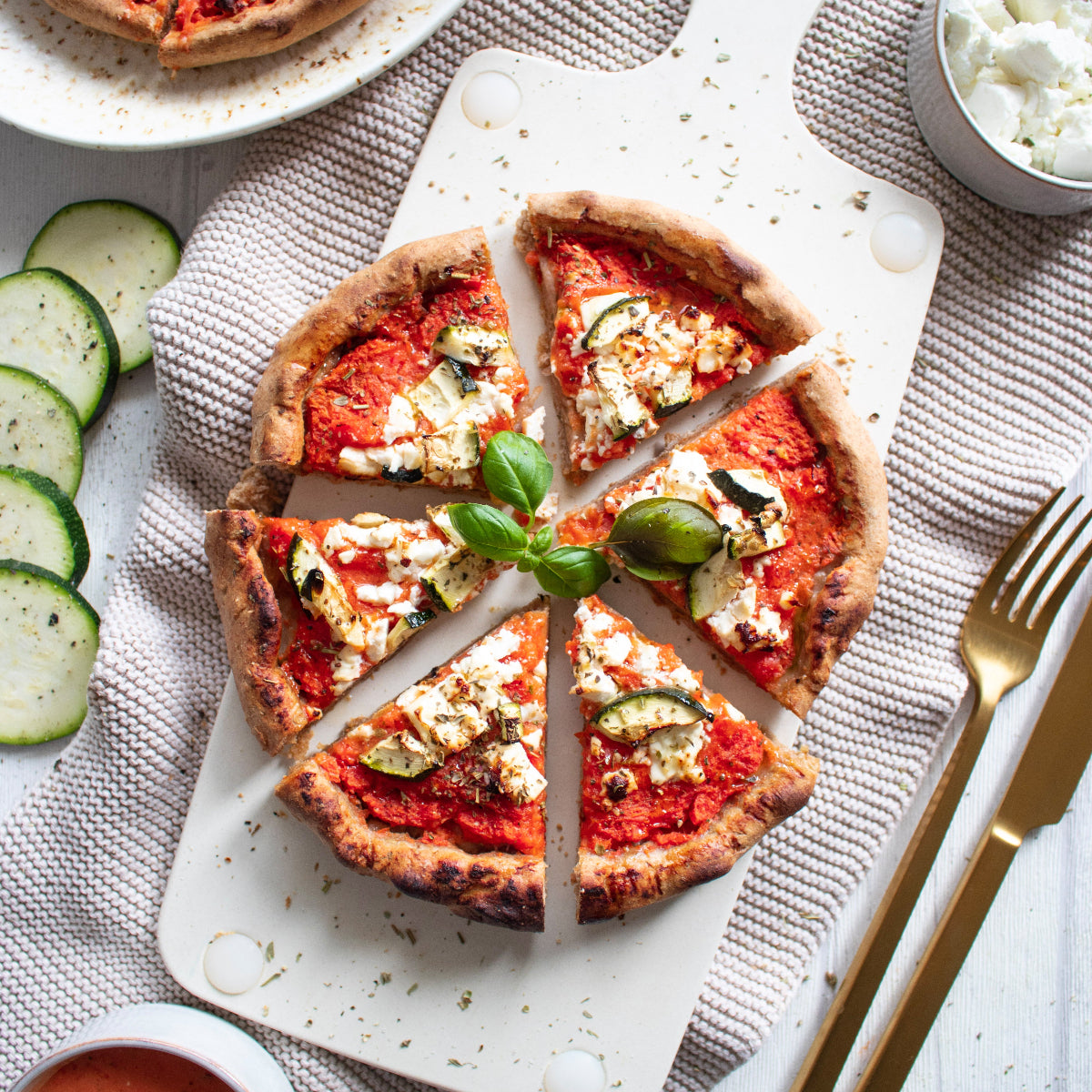 Produktbild der zubereiteten „Pizzaminikus“ Pizza mit knusprigem Rand, goldbraun gebacken. Belegt mit cremigem Hirtenkäse, Zucchinischeiben und frischem Basilikum auf fruchtiger Tomatensoße. Im Hintergrund eine Schale mit Feta, Zucchinischeiben und goldfarbenes Besteck auf heller Tischdecke.