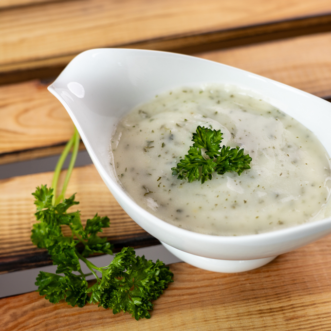 Weiße Sauciere mit cremiger Kräuter-Rahmsoße, garniert mit frischer Petersilie, auf einem Holzuntergrund angerichtet, daneben ein Petersilienzweig.