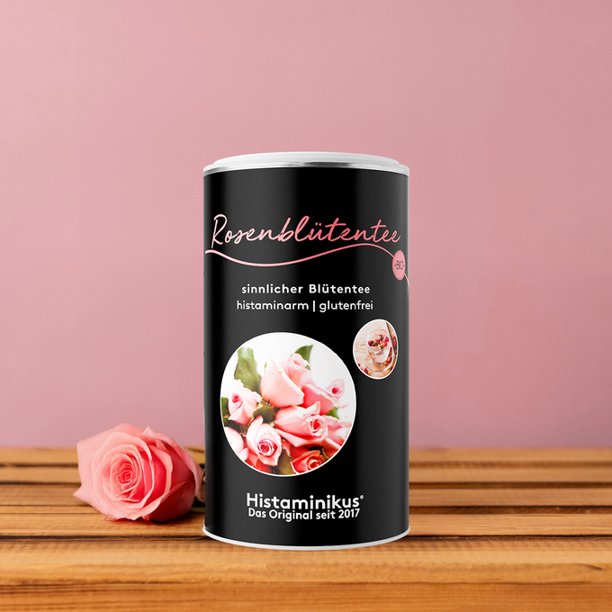 Schwarze Teedose „Rosenblütentee“ mit rosa Schrift und Abbildung einer Teetasse, dekoriert mit Rosenblüte und Minze vor rosafarbenem Hintergrund.