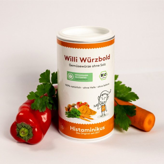 Weiße Gewürzdose mit orangefarbenem Etikett und der Aufschrift „Willi Würzbold – Gemüsewürze ohne Salz“. Daneben die Angaben „100 % natürlich“, „ohne Hefe“, „ohne Sellerie“. Darunter eine gezeichnete Figur mit Armbewegung sowie eine Illustration von Kurkuma, Karotten, Paprika, Ingwer und Kräutern. Links neben der Dose liegen eine rote Paprika und Petersilienblätter, rechts eine Karotte.