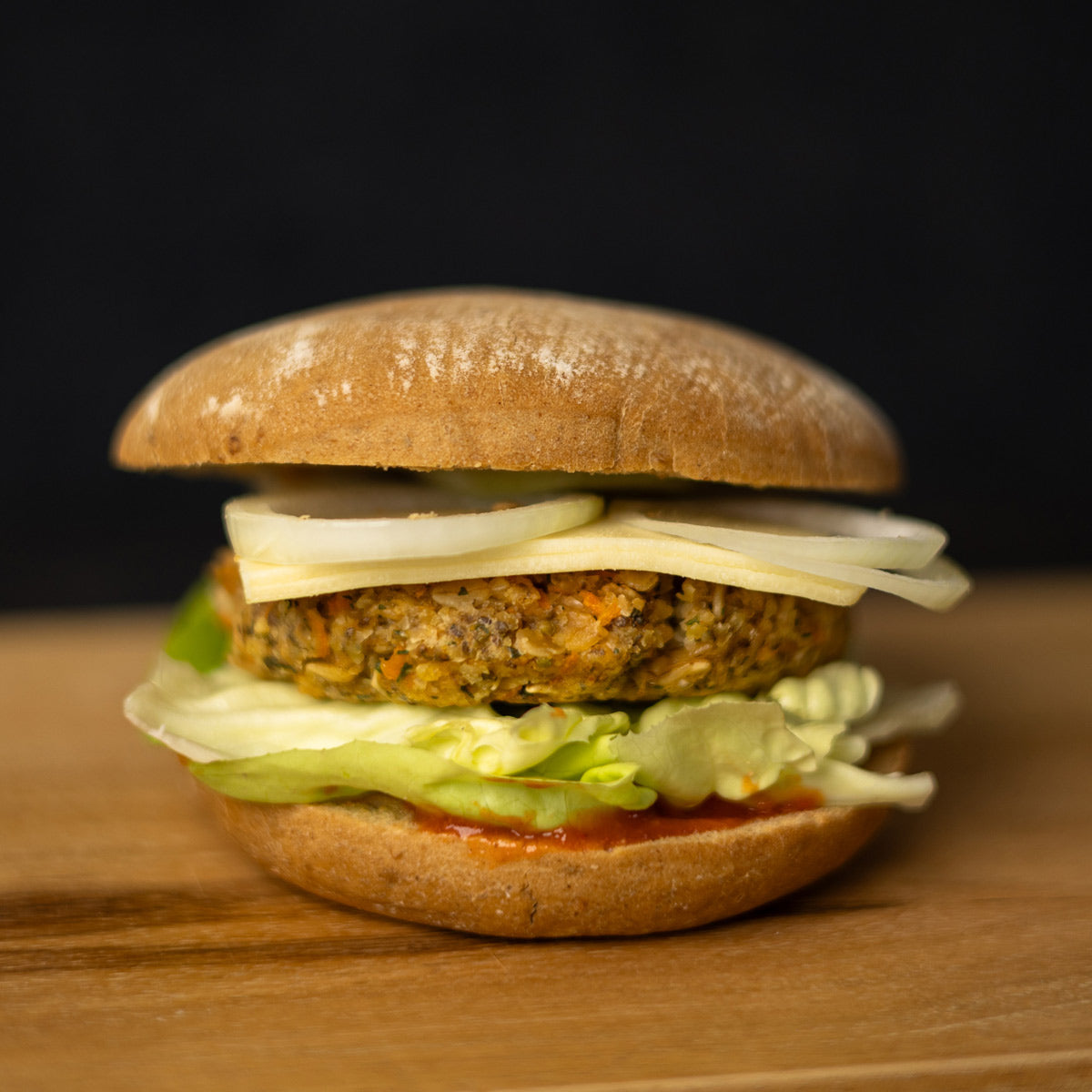 Burger mit runder Brötchenhälfte oben und unten. Zwischen den Brötchen ein pflanzlicher Patty mit sichtbaren Gemüseanteilen, darunter hellgrüner Blattsalat und rote Soße, darüber mehrere Scheiben roher Zwiebel. Der Burger steht auf einer Holzoberfläche, Hintergrund schwarz.