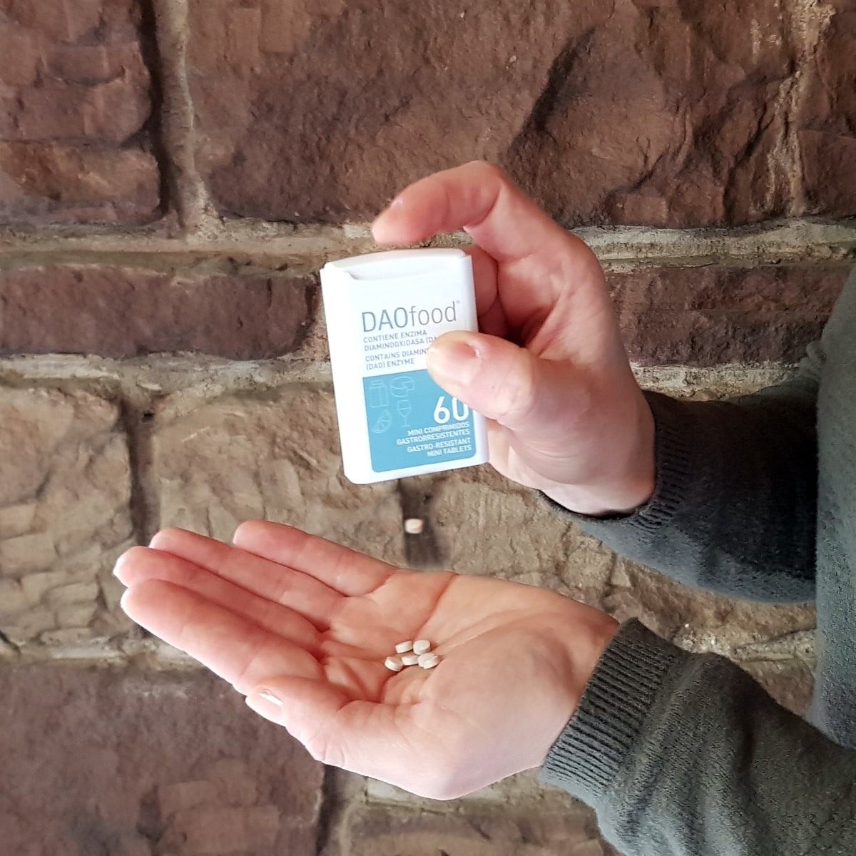 Eine Person hält einen DAOfood®-Spender in der rechten Hand und einige der kleinen Tabletten in der linken geöffneten Hand. Im Hintergrund eine grobe Steinmauer.