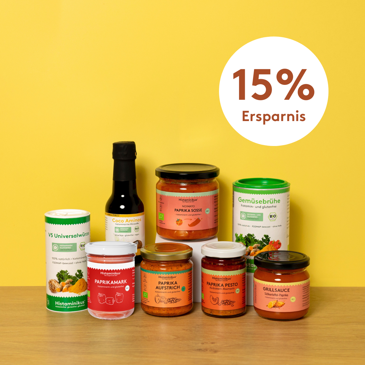 Acht histaminarme Bio-Produkte von Histaminikus auf hellem Holztisch vor gelbem Hintergrund: zwei Gewürzdosen, fünf Gläser mit Paprika-Aufstrichen, -Pesto, -Mark, -Soße und Grillsauce sowie eine Flasche Coco Aminos. Darüber ein Kreis mit rotem Schriftzug „15 % Ersparnis“.