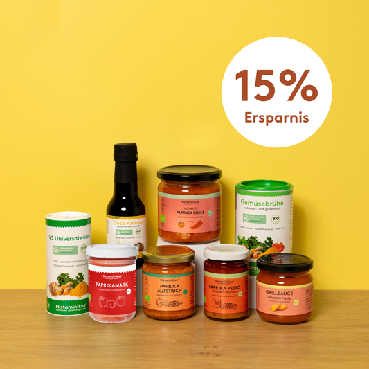 Acht histaminarme Bio-Produkte von Histaminikus auf hellem Holztisch vor gelbem Hintergrund: zwei Gewürzdosen, fünf Gläser mit Paprika-Aufstrichen, -Pesto, -Mark, -Soße und Grillsauce sowie eine Flasche Coco Aminos. Darüber ein Kreis mit rotem Schriftzug „15 % Ersparnis“.