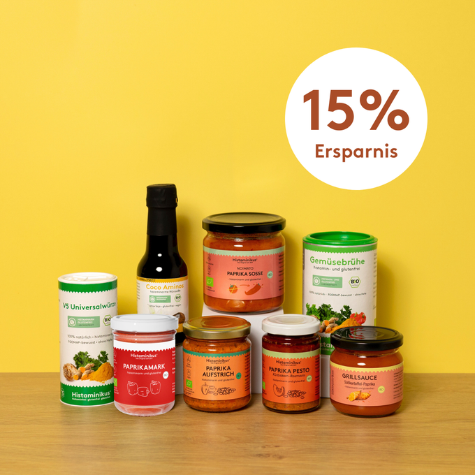 Acht histaminarme Bio-Produkte von Histaminikus auf hellem Holztisch vor gelbem Hintergrund: zwei Gewürzdosen, fünf Gläser mit Paprika-Aufstrichen, -Pesto, -Mark, -Soße und Grillsauce sowie eine Flasche Coco Aminos. Darüber ein Kreis mit rotem Schriftzug „15 % Ersparnis“.