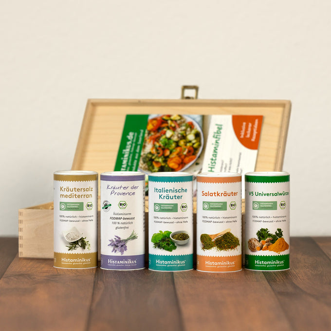 Fünf Gewürzdosen vor geöffneter Holzbox mit Rezeptheft im Hintergrund. Beschriftungen: „Kräutersalz mediterran“, „Kräuter der Provence“, „Italienische Kräuter“, „Salatkräuter“ und „V5 Universalwürze“. Jede Dose mit Bio-Siegel, Fotos der Kräuter und farblich passender Gestaltung.