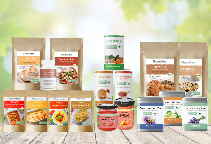 Verschiedene Histaminikus-Produkte auf einem Holztisch vor unscharfem, grünem Naturhintergrund: Backmischungen „Knuspriger Franz“ und „Pizzaminikus“, Gemüsebrühe, Currypulver, Paprikapulver, Müslimischungen „Hempies“ und „Basenmüsli“, drei Teedosen („Guten-Abend-Tee“, „Wohlfühltee“, „Entspannungstee“), L-Glutamin-Komplex, Grill- und Tomatensauce sowie vier Beutel „HistaFix“-Fertiggerichte.