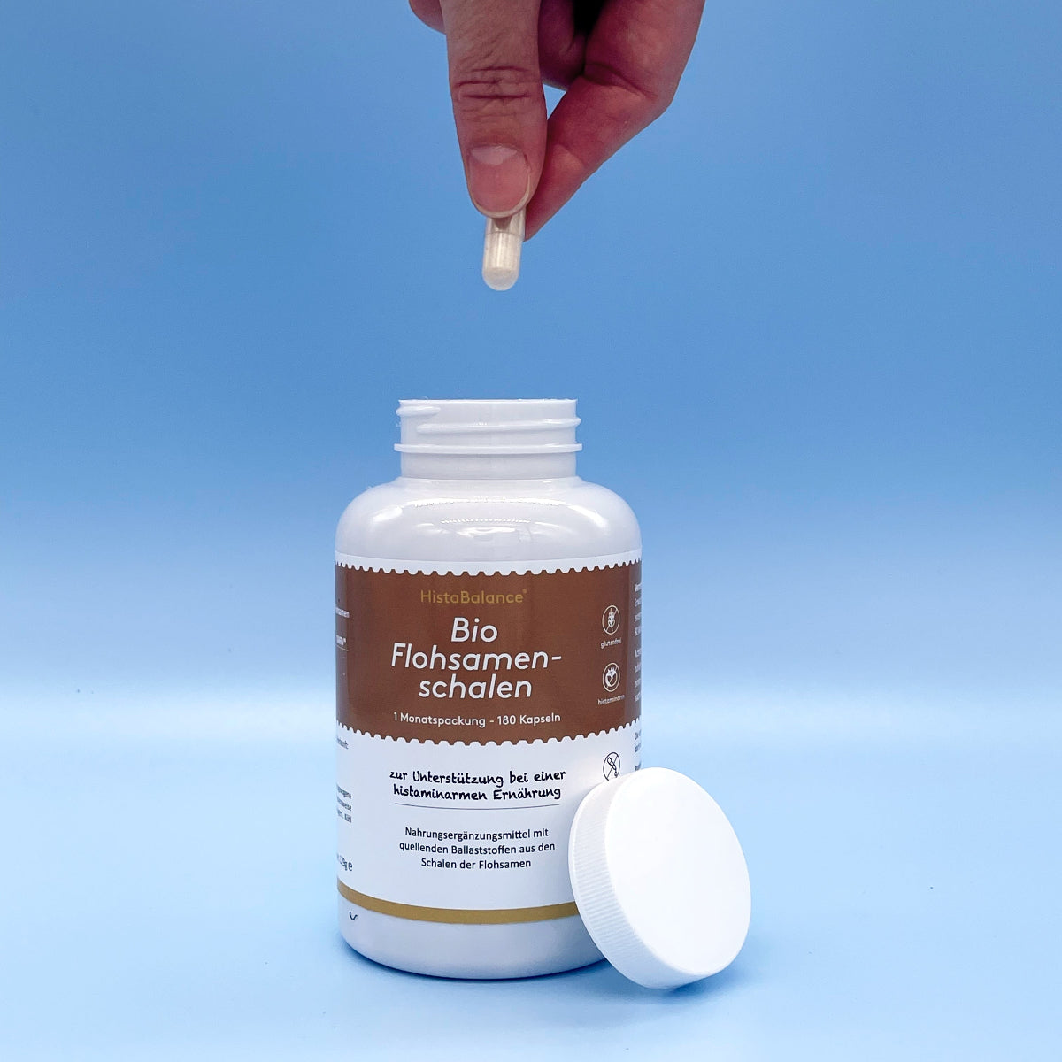 Dose „Bio Flohsamenschalen“ mit braunem Etikett auf blauem Hintergrund, Deckel abgenommen. Hand hält Kapsel über der geöffneten Dose.