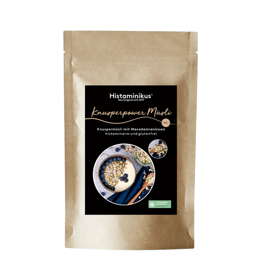 Produktfoto eines braunen Papierbeutels mit schwarzem Etikett. Darauf in weißer Schrift der Produktname „Knusperpower Müsli“ und die Beschreibung „Knuspermüsli mit Macadamianüssen – histaminarm und glutenfrei“. Abgebildet ist eine Müsli-Schale mit Joghurt und Blaubeeren.