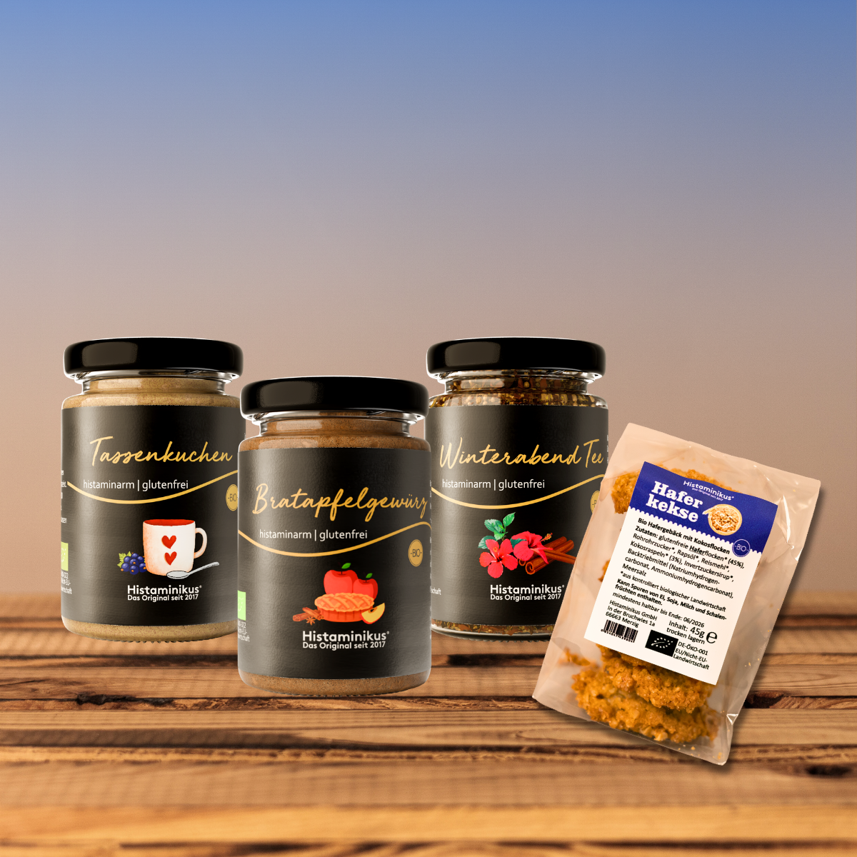 Vier histaminarme, glutenfreie Produkte von Histaminikus auf Holztisch: Tassenkuchen, Bratapfelgewürz und Winterabend Tee in Gläsern, daneben eine Packung Haferkekse, vor sanftem Farbverlauf-Hintergrund.