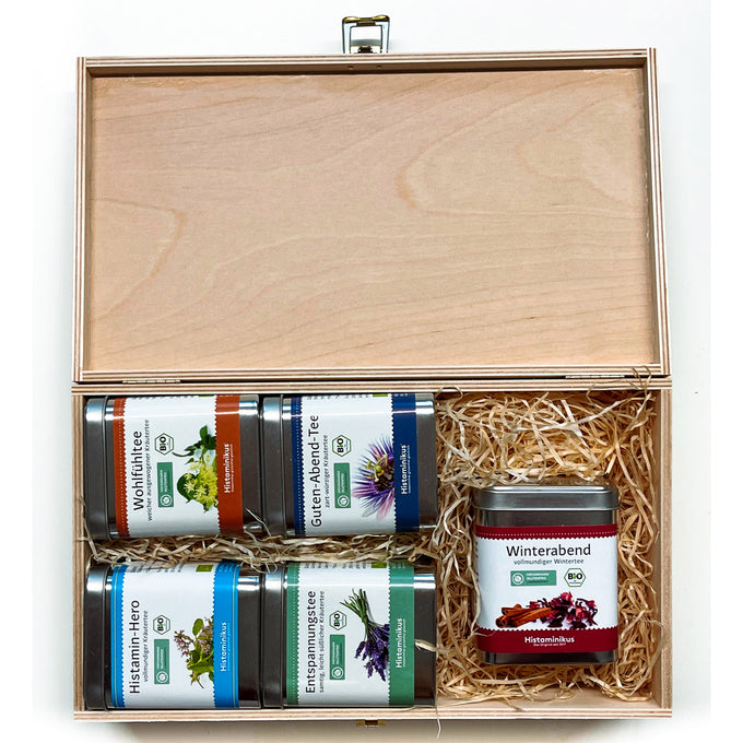 Offene Holzbox mit fünf Teedosen, eingebettet in Holzwolle. Sorten: „Wohlfühltee“, „Guten-Abend-Tee“, „Histamin-Hero“, „Entspannungstee“ und rechts separat platziert „Winterabend“. Jede Dose mit farbigem Etikett, Kräutermotiv und Bio-Siegel.