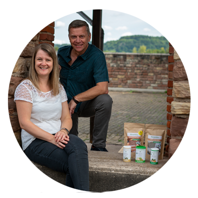 Thomas und Michaela Zinser, Gründer von Histaminikus, sitzen lächelnd nebeneinander auf einer Steinstufe vor einer Backsteinmauer mit Blick auf eine Landschaft. Neben ihnen stehen mehrere Histaminikus-Produkte, darunter Gewürzdosen und Papiertüten.