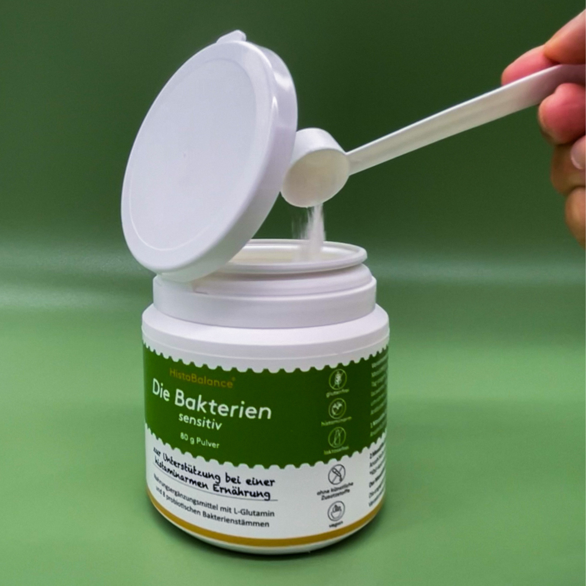 Offene Dose „Die Bakterien sensitiv“ mit grünem Etikett. Eine Hand füllt mithilfe eines weißen Dosierlöffels Pulver aus der Dose.