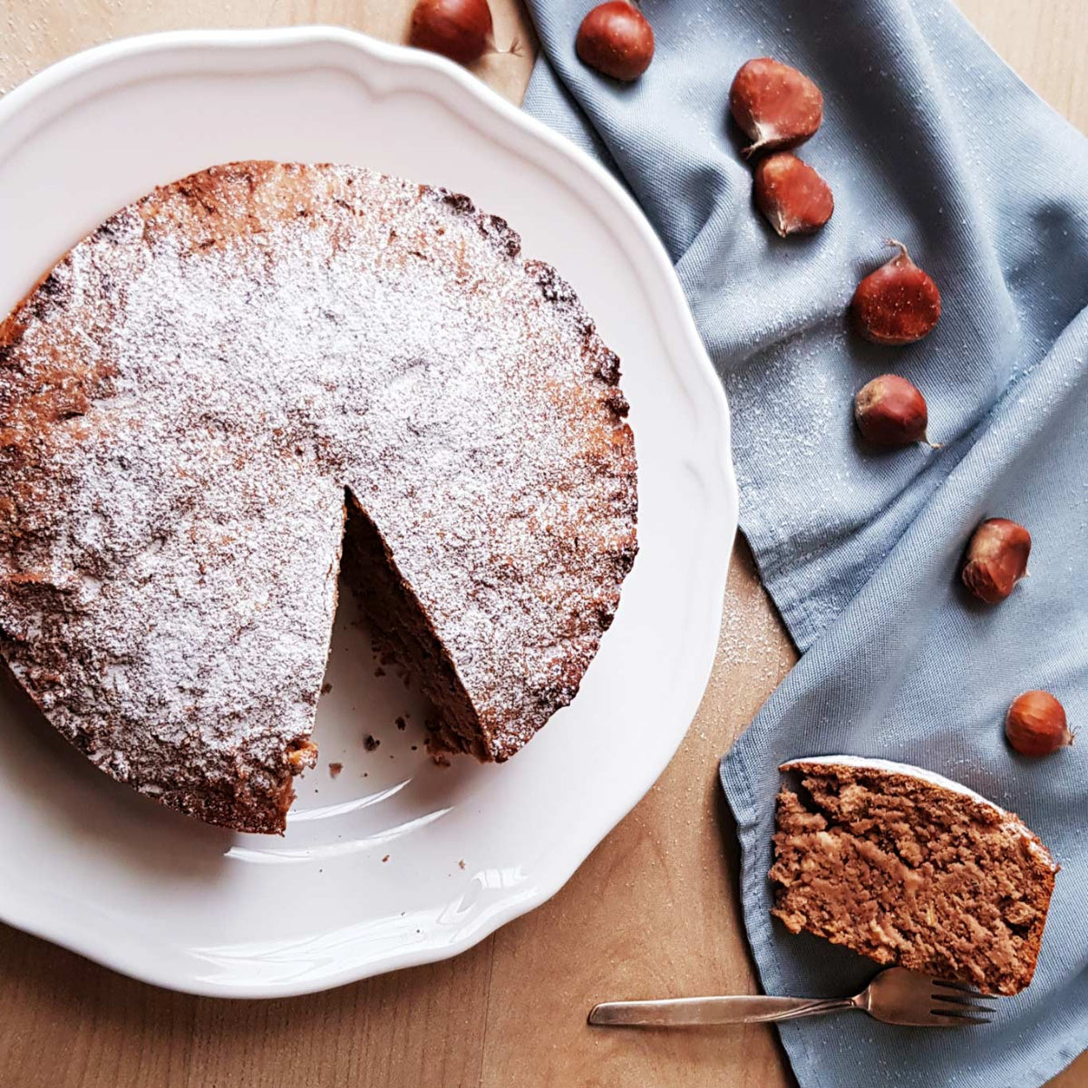 Runder Kuchen mit Puderzucker bestäubt auf weißem Teller, ein Stück herausgeschnitten. Daneben braune Haselnüsse auf blauer Stoffserviette.